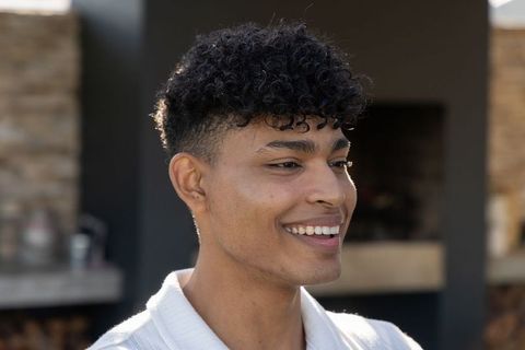 African american man smiling and looking right wearing white knit top on backyard patio with hearth