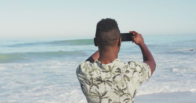 Rear view of african american man with smartphone on sunny beach taking photos of the sea. Summer, free time, relaxation, communication and vacations.
