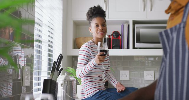 Image of happy african american couple drinking wine and talking in kitchen. love, relationship, togetherness and spending quality time at home.