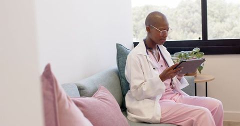 Calm medical professional using tablet on pastel clinic sofa wearing pink scrubs and coat