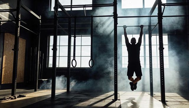 Silhouetted athlete hanging from pull-up bar in foggy industrial gym with rig rings