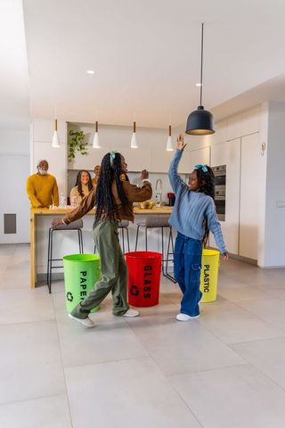 Sisters dancing by color-coded recycling bins in modern open-plan kitchen with family