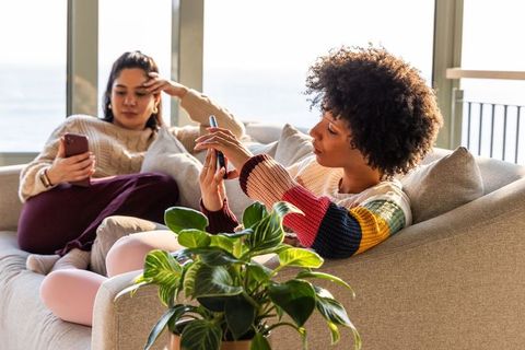 Diverse female friends lounging on sofa scrolling smartphones in bright modern apartment