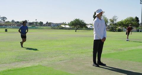 Standing coach watching community sports practice on sunny field wearing white bucket hat