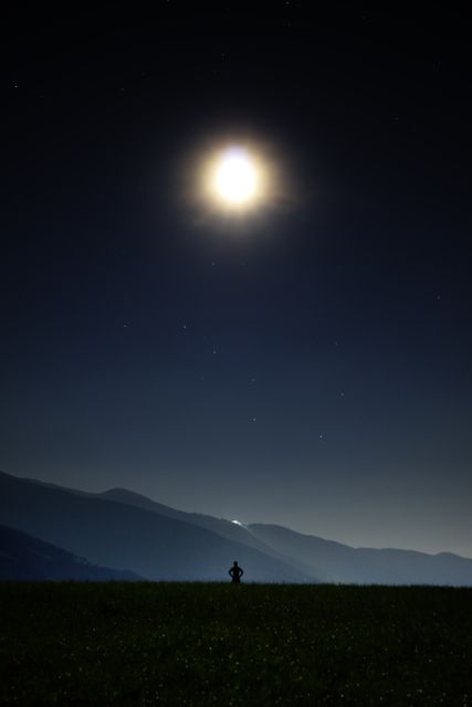Person standing on open field under moonlit sky with mountain ranges in background; perfect for themes of solitude, serenity, nature, night, astrophotography, and inspiration. Excellent for travel brochures, nature-focused products, meditation guides, and inspirational content.