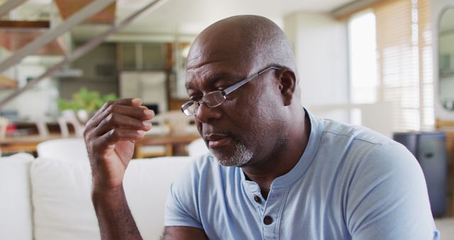 African American man sitting at home appearing thoughtful and introspective. Wearing glasses and casual blue shirt, located in modern living room, suitable for depicting contemplation, retrospection, mental health awareness, or moments of personal reflection. Ideal for use in articles or content about mental health, lifestyle, midlife introspection, or everyday living.