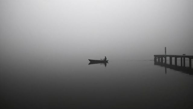 Solitary rower gliding through dense morning fog on glassy lake near wooden jetty