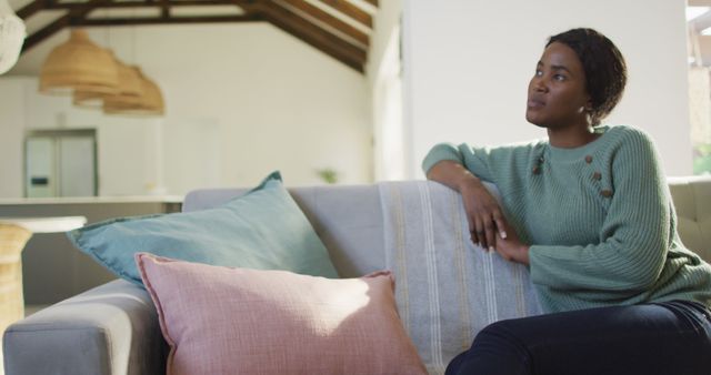 Woman cozy on couch, modern living room, thoughtful. Ideal for articles on relaxation, home interior, and self-care. Great for advertisements on furniture, home design, or lifestyle brands. Perfect for blogs emphasizing personal time, mental health, or cozy environments.