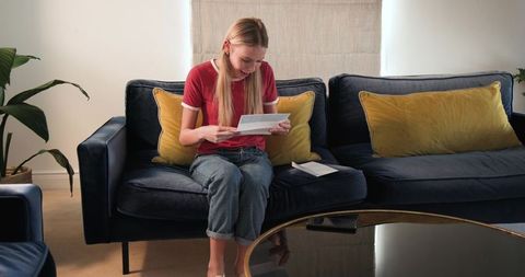 Young woman reading letter on navy sofa in cozy modern living room, smiling