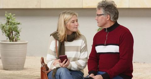 Senior couple sitting on ledge holding smartphones sharing conversation in urban plaza