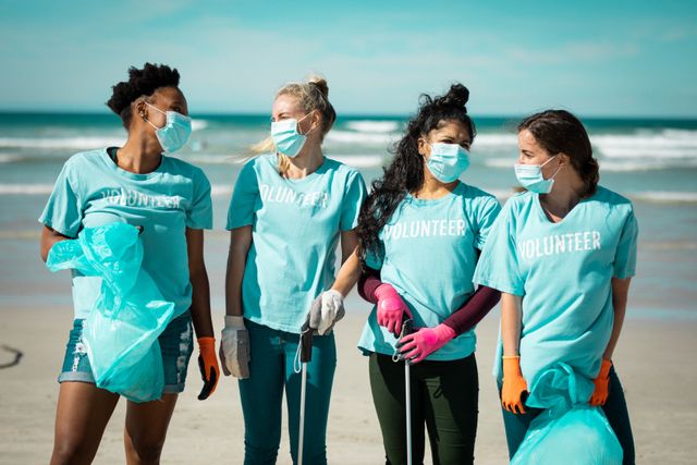 Diverse group of women wearing volunteer t shirts and face masks picking up rubbish from beach. eco conservation volunteers, beach clean-up during coronavirus covid 19 pandemic.