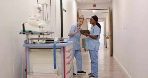Reviewing tablet chart, nurses collaborating in hospital corridor with crash cart