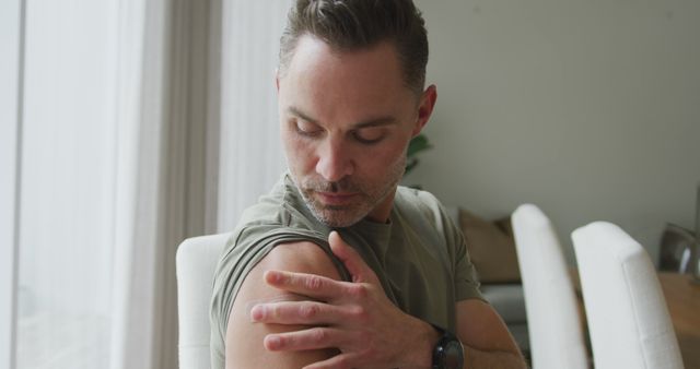 Man looking at his arm while sitting indoors in a modern living room with natural light. Useful for campaigns focused on health and wellness, self-care, medical check-ups or home-inspired lifestyles. Suitable for use in blogs, articles, and promotional materials related to personal health awareness.
