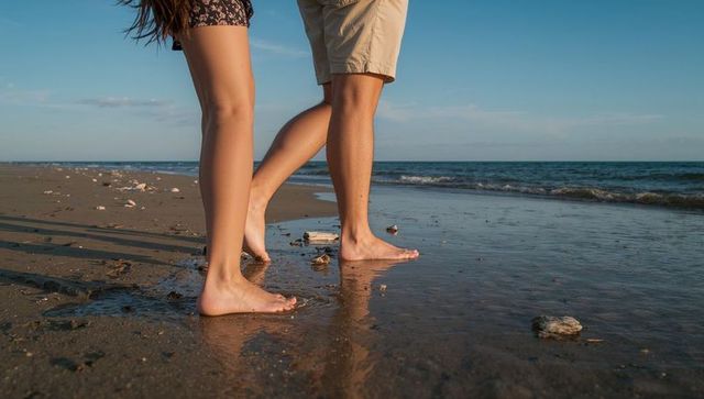 Couple walking barefoot on wet sand at sunset, legs and reflections in shallow water