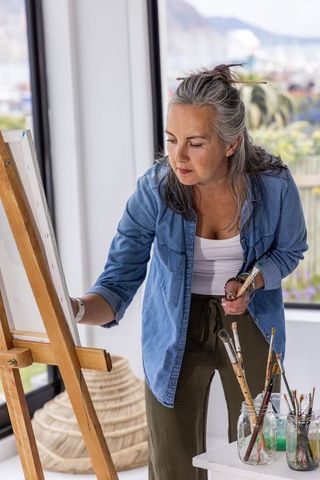 Middle-aged woman painting on easel in sunlit coastal studio overlooking harbor