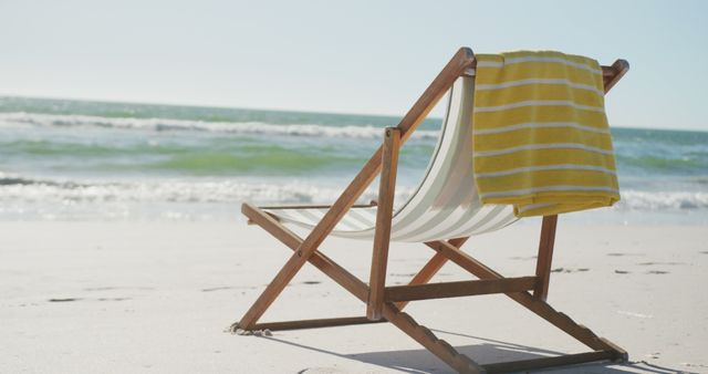 A wooden beach lounger with a striped yellow towel is lying on a sandy beach under clear skies. Ideal for promoting travel destinations, vacation rentals, summer activities, and lifestyle blogs focusing on relaxation and leisure time.