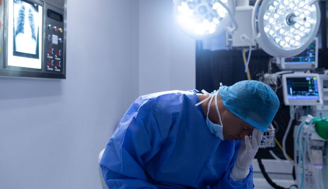 Surgeon sitting in an operation theater, appearing tense with hand on forehead. Medical equipment and x-rays visible. Ideal for use in articles or materials related to high-stress medical professions, healthcare challenges, surgeon mental health, or workplace stress.