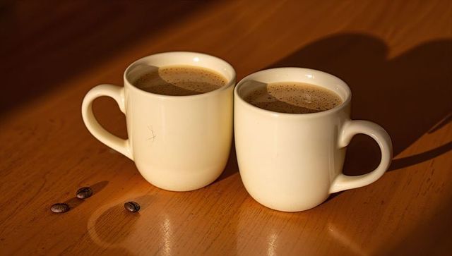 Two white ceramic coffee mugs with crema on sunlit wooden table, steaming morning cups