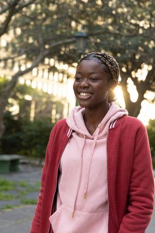 Smiling teen girl on park path during golden hour wearing pink hoodie and red jacket