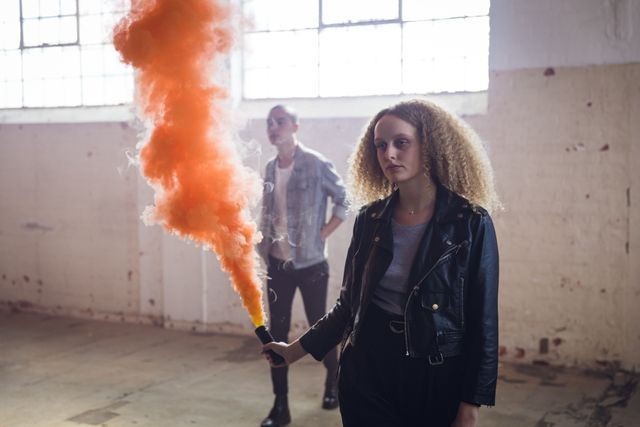 Young woman with curly hair holding an orange smoke grenade in an empty warehouse, creating an edgy and urban atmosphere. A young man stands in the background, adding to the alternative and modern vibe. Ideal for use in fashion editorials, urban lifestyle blogs, and promotional materials for edgy brands.