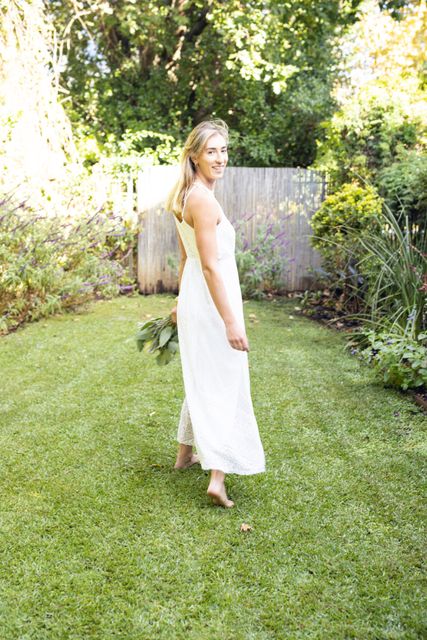 Portrait of smiling caucasian young woman wearing white dress walking on lush grassy field in yard. Unaltered, lifestyle, happy and nature concept.