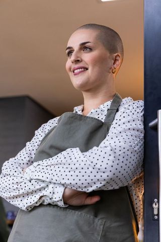 Confident cafe owner with shaved head wearing apron and polka dot blouse at doorway smiling