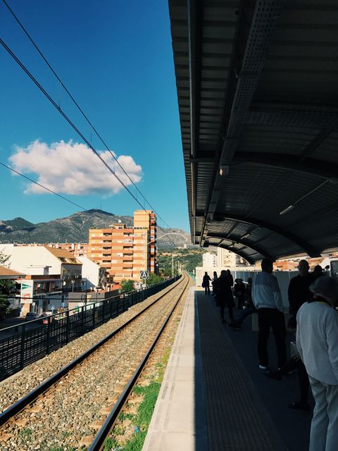 People standing on a train platform under a covered area. Bright sunny day with clear blue sky and scenic mountain in background. Urban buildings and railway tracks visible, creating a blend of natural and urban scenery. Ideal for projects involving transportation, travel, city life, commuting scenes, and scenic railway journeys.