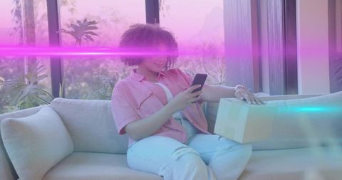 Curly-haired woman relaxing on sofa using smartphone while reaching for parcel