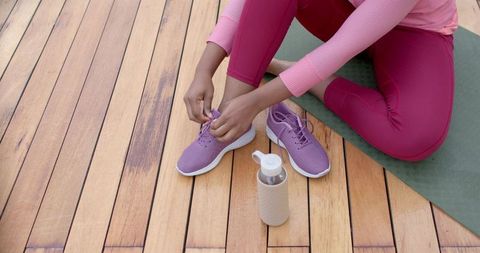 African american woman tying purple running shoe on yoga mat on wooden deck for workout