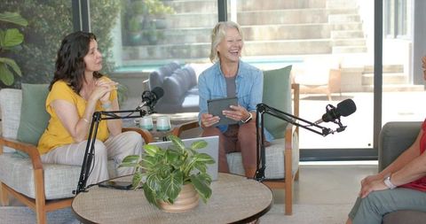 Women hosting cozy living room podcast laughing over tablet and microphones
