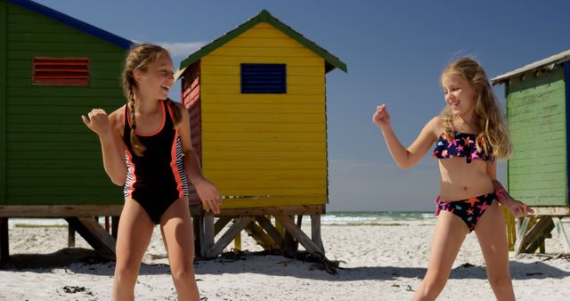 Happy caucasian girls in swimsuits dancing and smiling near wooden houses on sunny beach. Childhood, friendship, free time, summer, travel, vacations and lifestyle, unaltered.