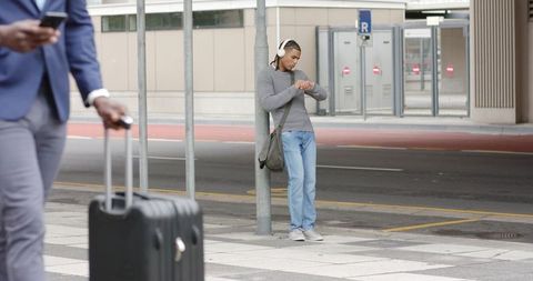 Diverse commuters waiting at curb, man with headphones checking watch, businessman pulling luggage