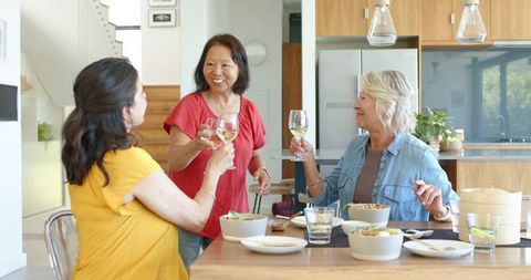 Mature women clinking wine glasses over casual lunch in bright modern kitchen