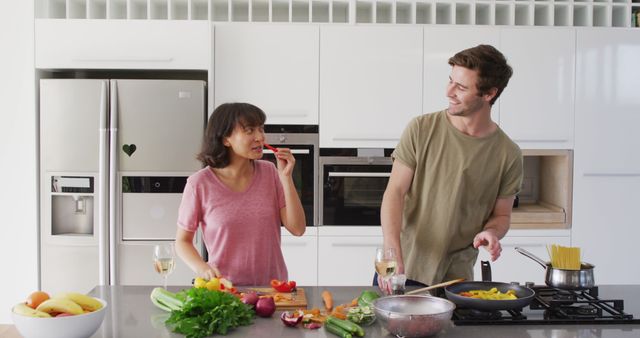 Couple enjoying time cooking together in a modern kitchen. Both are handling different tasks - chopping vegetables and stirring noodles in a pan - while smiling and bonding. Perfect for illustrating themes of domestic life, relationship bonding, healthy living, and modern home interiors.
