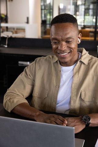 African American man working on laptop in modern open-plan office wearing wireless earbuds
