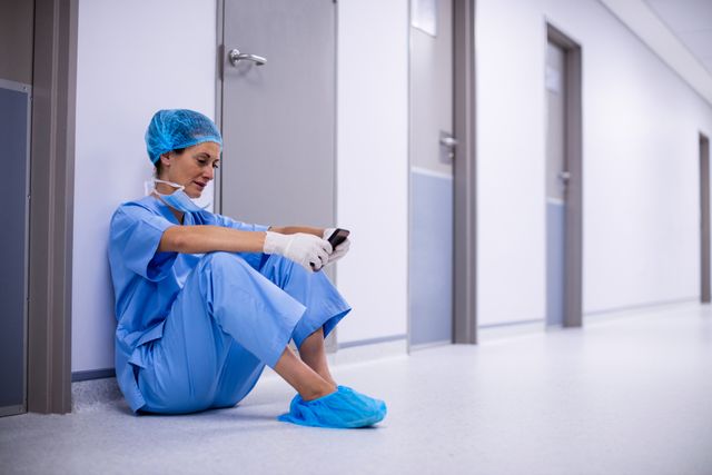 Surgeon in blue scrubs and protective gear sitting on hospital floor using mobile phone. Ideal for depicting healthcare professionals taking a break, the use of technology in healthcare, or the human side of medical workers. Useful for articles, blogs, and advertisements related to healthcare, medical professions, and hospital environments.