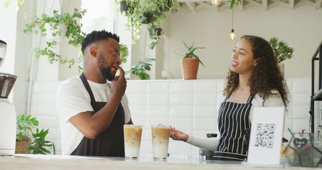 Two baristas having discussion at the counter inside a brightly lit coffee shop. Iced coffee drinks on countertop signify focus on customer service. Can be used for themes related to teamwork, cafe culture, hospitality industry, and small business operations.