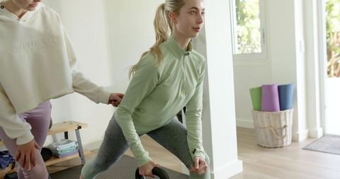 Two women lunging with kettlebell in sunlit home gym, one spotting for safe strength training