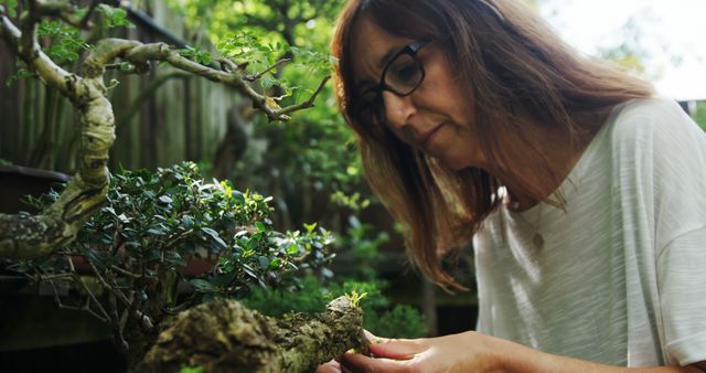Woman delicately pruning a bonsai tree in a tranquil garden setting. Ideal for content related to gardening, relaxation techniques, hobbies, meticulous craftsmanship, outdoor activities, and nature appreciation.