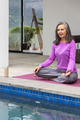 Senior woman meditating on poolside yoga mat in purple top promoting calm wellness