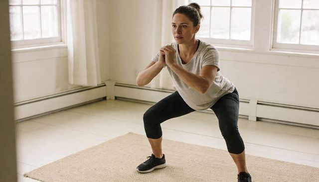 Woman performing home squat workout in bright minimalist room with woven rug and sneakers