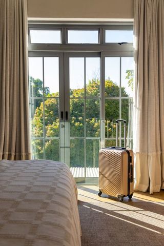 Tan hard-shell rolling suitcase standing by sunlit french doors with balcony view