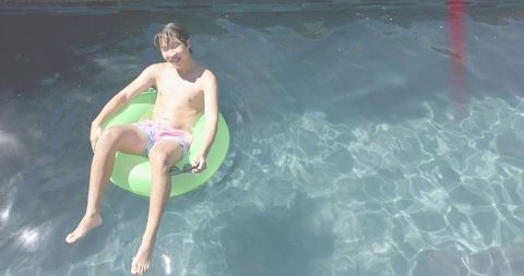 Asian man relaxing on green inflatable ring in sunlit backyard pool wearing trunks