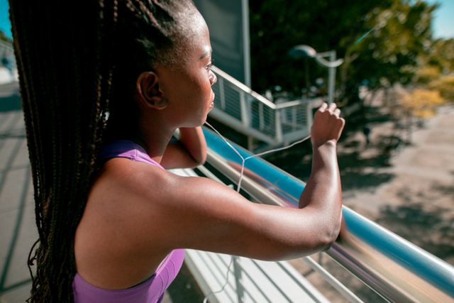 African American woman wearing earphones and sportswear, resting on a bridge at sunset. Ideal for promoting healthy lifestyles, fitness routines, outdoor activities, and urban exercise. Can be used in advertisements, fitness blogs, and wellness campaigns.