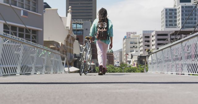Rear view of a biracial man with long dreadlocks out and about in the city on a sunny day, wearing backpack, walking the street and wheeling his bicycle in slow motion.