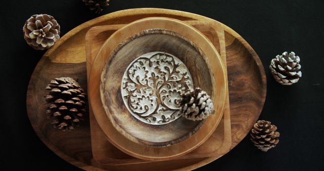A wooden plate with an intricate design is centered on a dark background, surrounded by pine cones and smaller wooden dishes. Its rustic charm and natural elements suggest a cozy, earthy aesthetic ideal for home decor enthusiasts.
