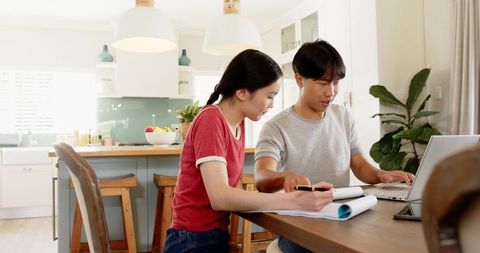 Asian Couple Collaborating at Kitchen Table with Laptop and Notebook