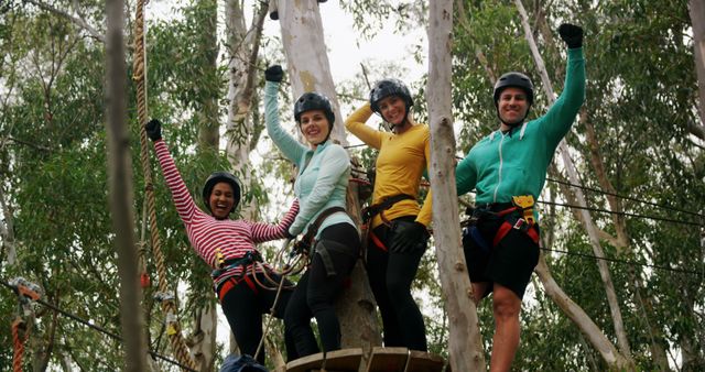 Group of smiling friends in colorful outfits enjoying a thrilling zip line adventure high among trees in a forest. Wearing safety helmets and harnesses, they display excitement and celebration. Perfect for depicting outdoor activities, teamwork, adventure tourism, promotional materials for adventure parks or team-building workshops.