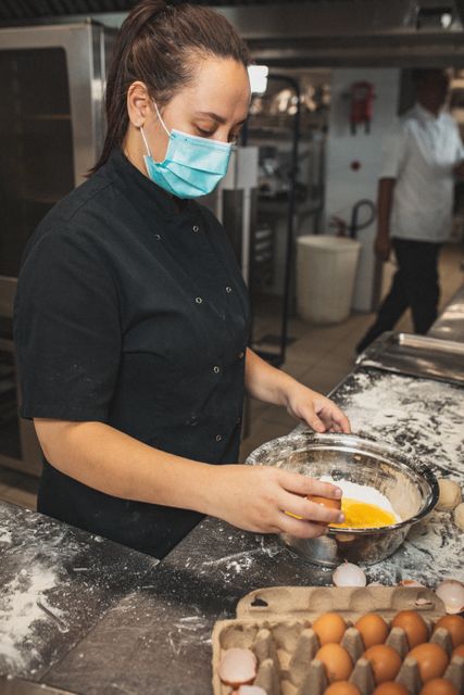Female chef wearing a mask preparing a meal with eggs in a busy restaurant kitchen. Ideal for use in articles or advertisements related to culinary arts, professional cooking, restaurant industry, food safety, and hygiene practices during the COVID-19 pandemic.