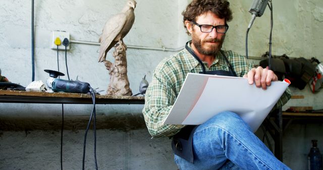 Sculptor sitting in workshop, deeply concentrated, studying a sketch for his next sculpture project. Workspace is filled with tools and an unfinished sculpture of a bird, reflecting creativity and dedication to craftsmanship. Ideal for use in articles or ads about art, creativity, craftsmanship, and artistic professions.